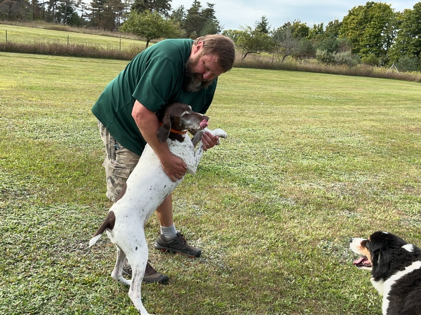Jonathan hugging a pointer who is standing up on its hind legs for the hug, while a fluffy puppy watches