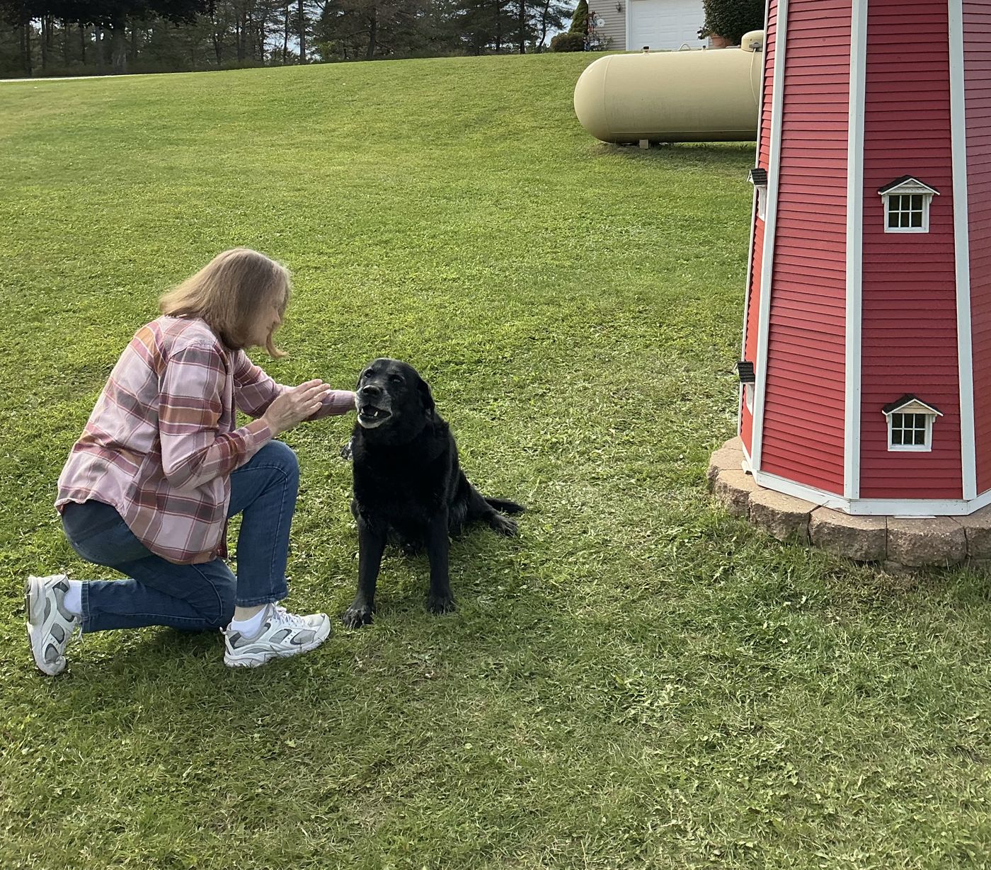 Sharon kneeling in the grass to greet a gray-muzzled black Lab beside the red lighthouse in the yard